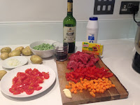 Ingredienti per spezzatino: carne di manzo, patate, pomodorini, piselli, vino rosso, carote, cipolla, aglio e brodo di pollo, disposti su un piano di lavoro.