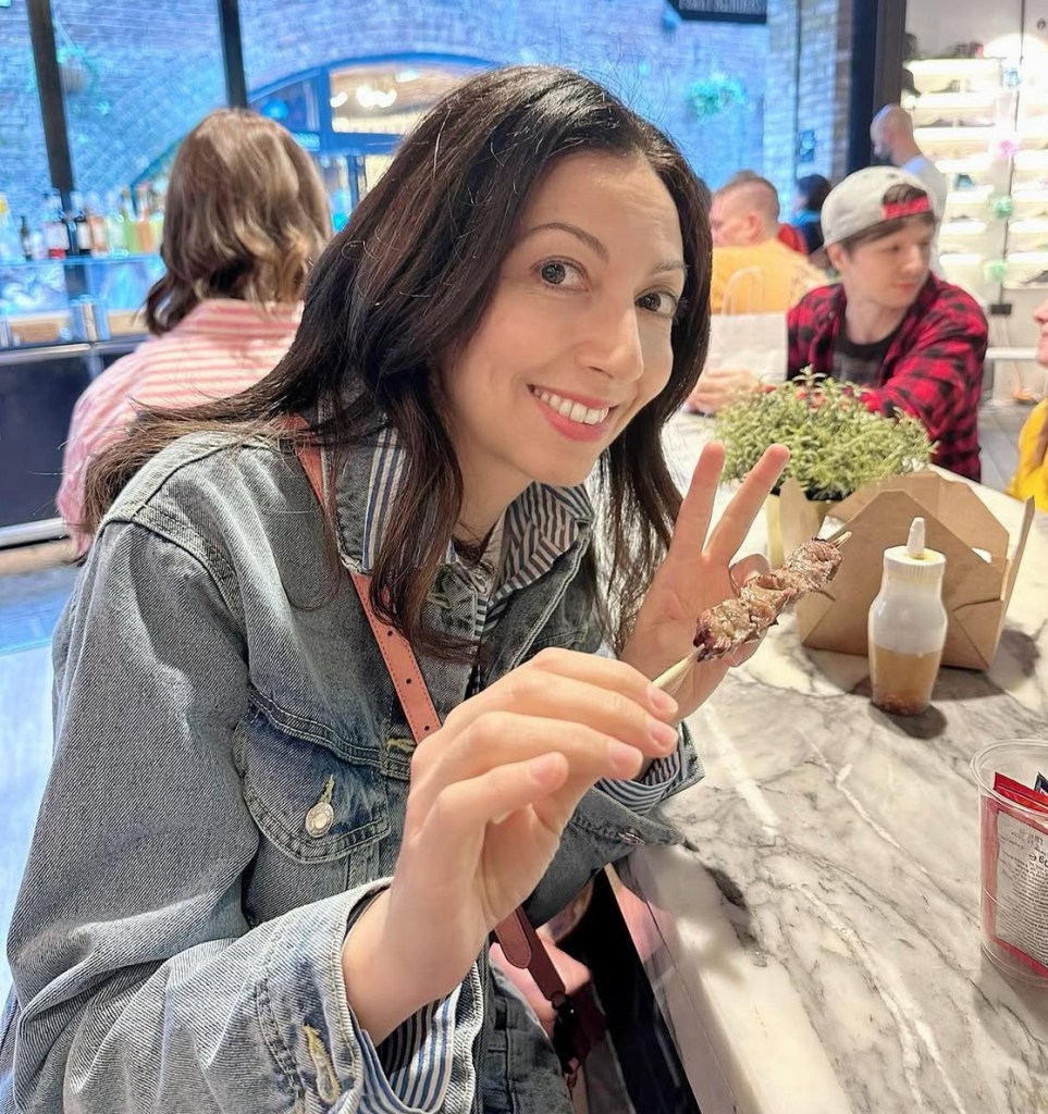 Persona sorridente che tiene in mano un arrosticino, seduta a un tavolo in un locale vivace.