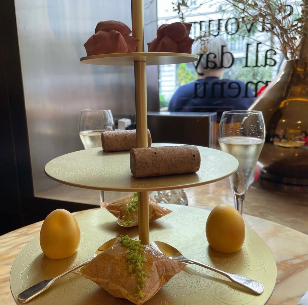 Un elegante vassoio per afternoon tea con dolci creativi, tra cui due dolcetti di pasta rosa, rotoli di cioccolato e un uovo di dessert, accompagnato da un bicchiere di champagne.Da Cakes and bubbles a Londra