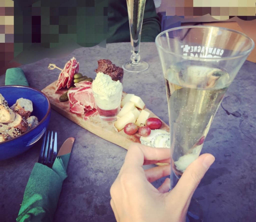 Un bicchiere di champagne in primo piano, accompagnato da un tagliere con formaggi, salumi e sottaceti su una tavola. Sullo sfondo, una ciotola di pane rustico e posate. Bistrot Champagne + Fromage a Londra.