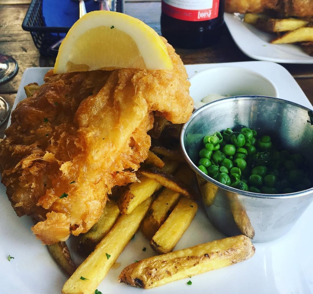Piatto di fish and chips con filetto di pesce fritto croccante, patatine fritte e una porzione di piselli verdi, decorato con una fetta di limone.