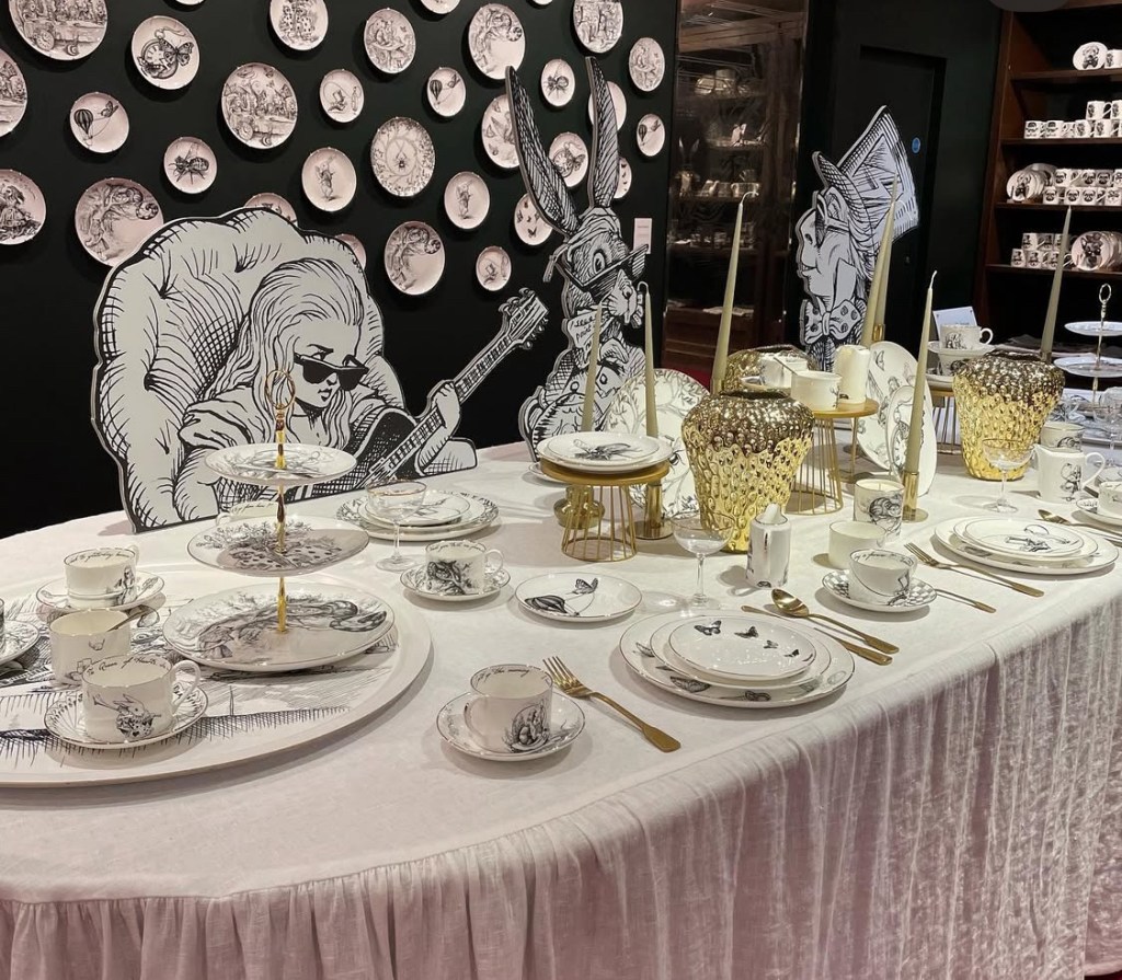 Un tavolo imbandito per l'afternoon tea, decorato con porcellane artistiche e illuminato da candelabri dorati, con illustrazioni in bianco e nero sullo sfondo.Da Fortnum Mason a Londra