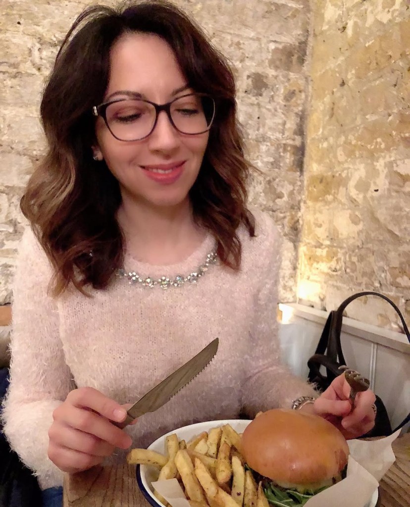 Una persona sorridente che tiene un hamburger e delle patatine fritte, seduta in un ambiente accogliente con pareti di pietra da Honest Burger Londra.