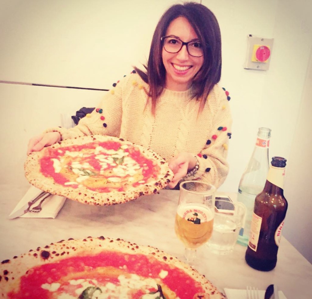 Una persona sorridente tiene in mano una grande pizza con salsa di pomodoro e mozzarella. Sul tavolo ci sono anche delle bevande.Pizza napoletana a Londra
