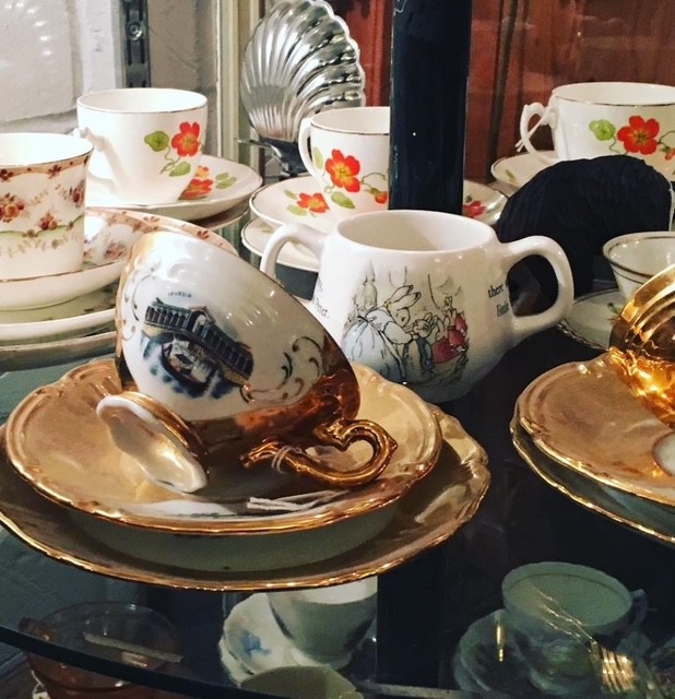 Un'esposizione di tazze e piattini in porcellana, tra cui una tazza decorata con dettagli dorati e una porzione di tè. In background, si intravedono diverse altre tazze con motivi floreali.