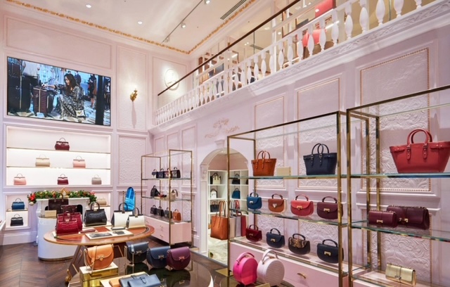 Interno elegante di una boutique di borse con pareti rosa e scaffali in vetro. Diverse borse di design sono esposte, mentre uno schermo nella parte superiore mostra un'immagine di modella che prova accessori.