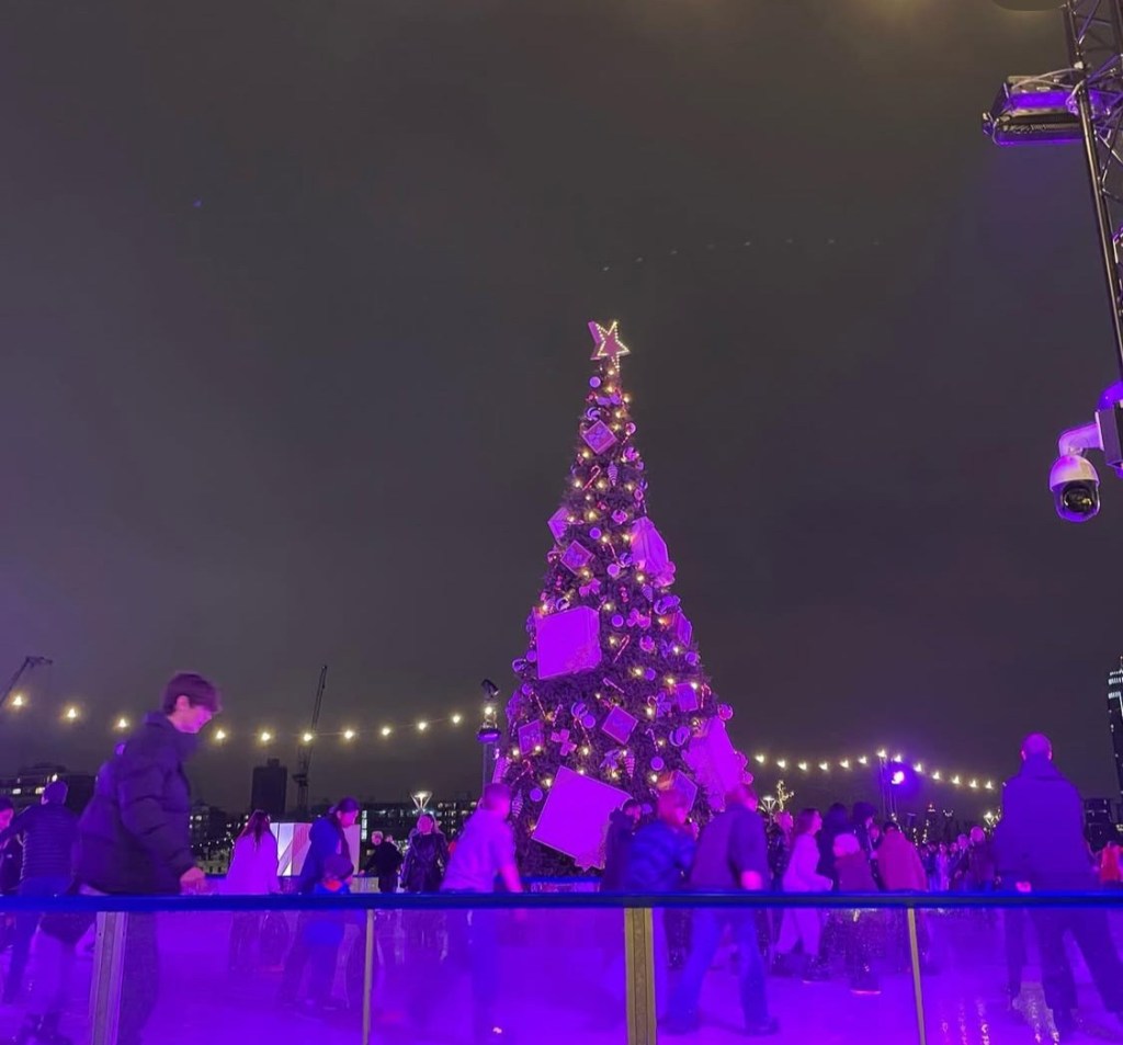 Un albero di Natale decorato con luci e regali, circondato da persone che pattinano su una pista di ghiaccio al chiaro di luna.