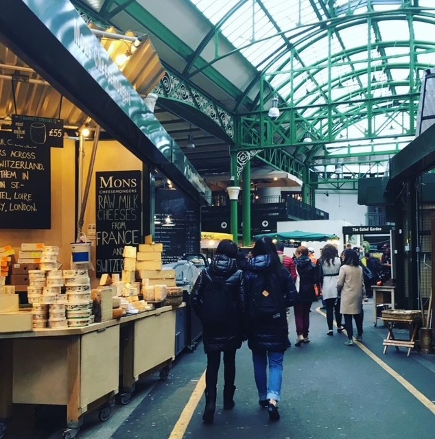 Un mercato di Londra con bancarelle allestite di formaggi e cibo, mentre visitatori passeggiano sotto una struttura in vetro e metallo.