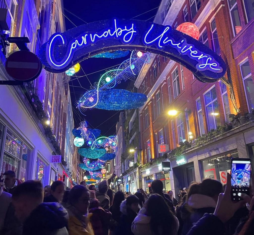 Una strada di Carnaby Street a Londra decorata con luci natalizie luminose e il grande cartello "Carnaby Universe", circondata da una folla di persone che passeggiano sotto le luminarie.