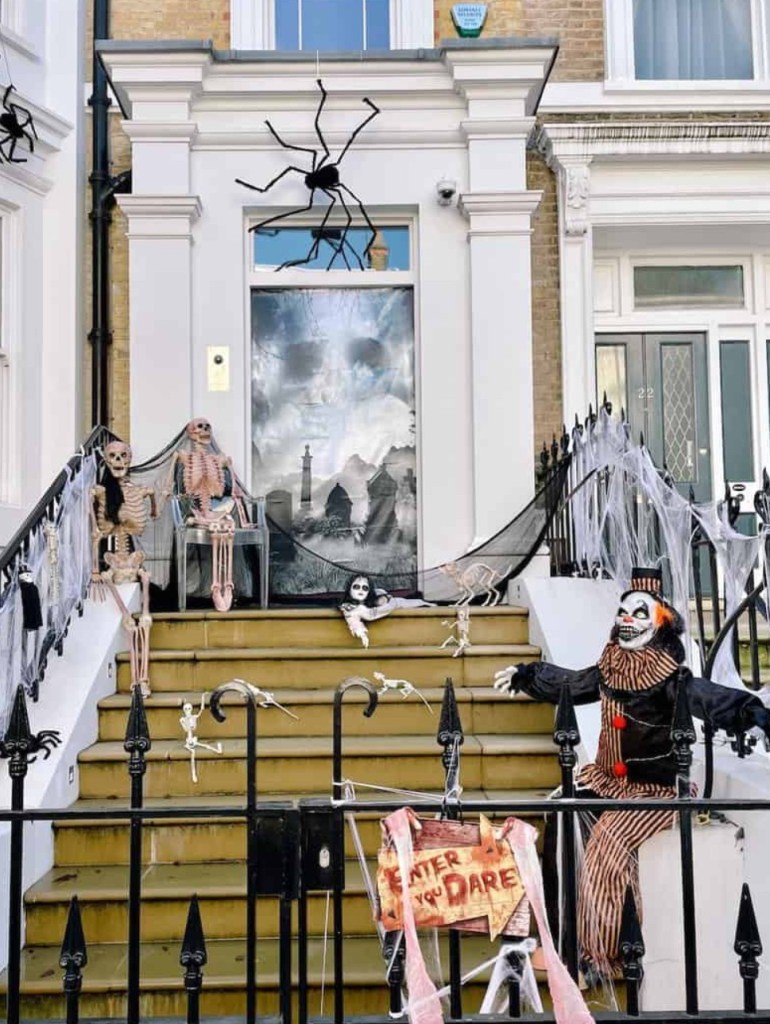 Decorazioni di Halloween a Londra su una scala, con scheletri, ragnatele e un clown spaventoso, accanto a un cartello che dice 'Enter if you dare'.