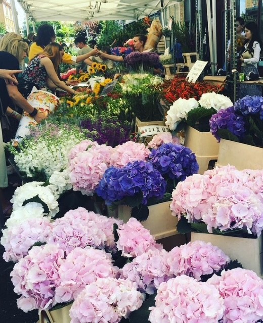 Un mercato di fiori colorati con diverse varietà di fiori freschi, tra cui grandi mazzi di ortensie rosa e blu, mentre alcune persone scelgono i fiori dai banchi.