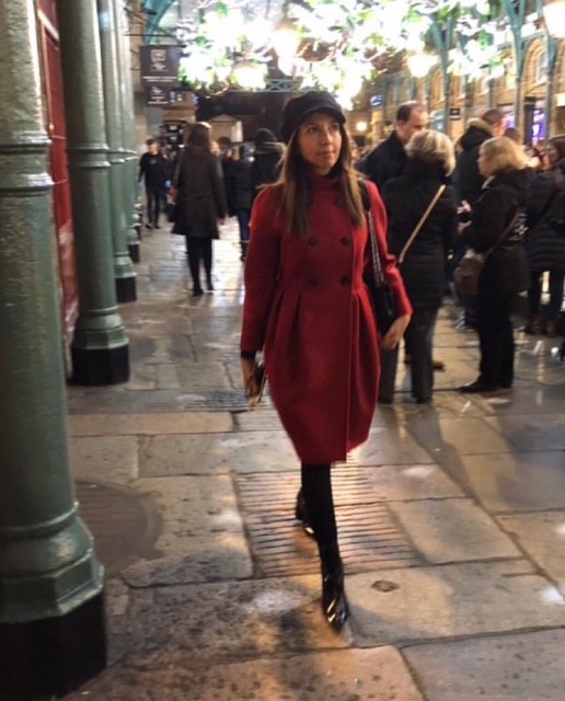 Una persona in un cappotto rosso cammina in un mercato affollato, circondata da altri visitatori e colonne verdi.