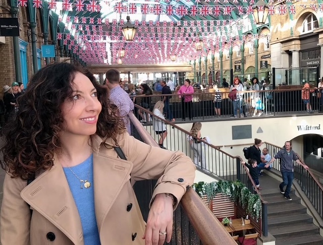 Una persona con capelli ricci e scuri indossa un trench beige mentre osserva un mercato affollato decorato con bandierine britanniche.