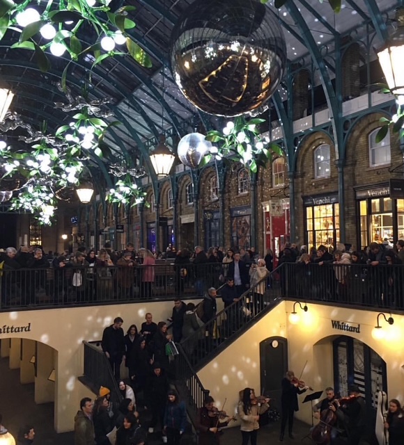 Interno di un mercato con decorazioni festive, luci appese e una folla di persone che ascoltano musicisti di strada.