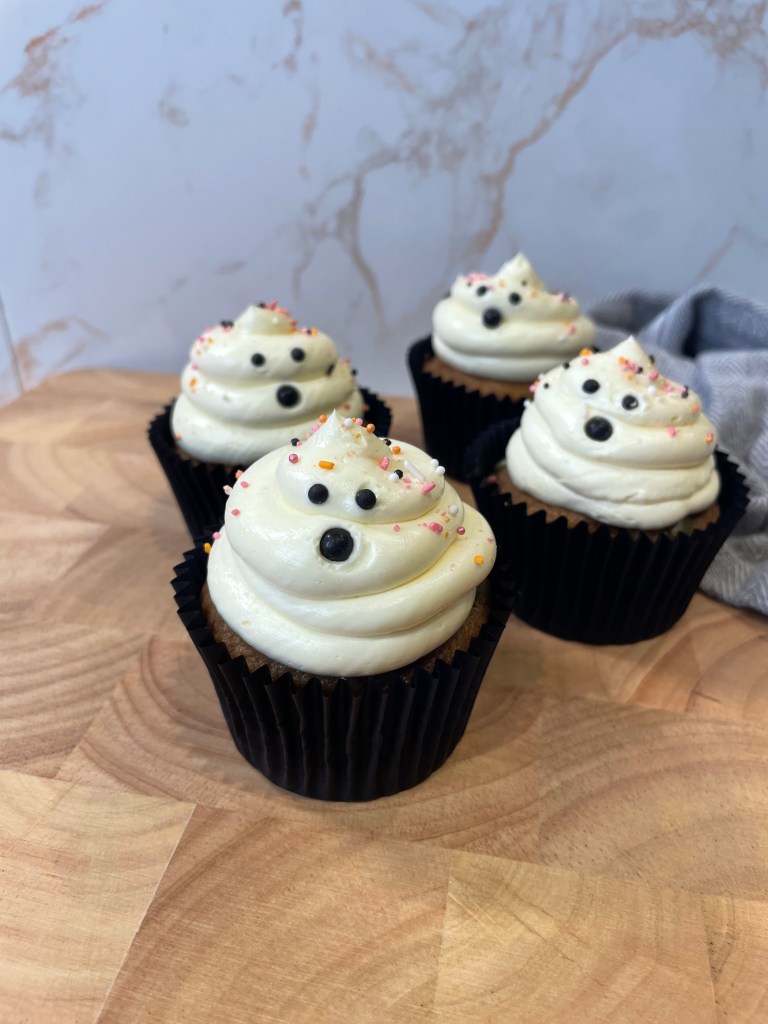 Cupcake decorato con crema di formaggio bianco, guarnito con perline colorate e uno sfondo di legno chiaro.