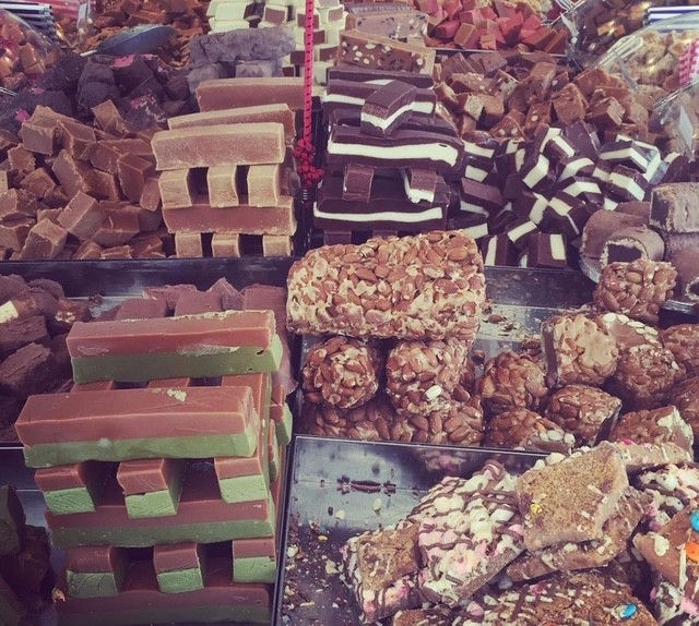 Un assortimento colorato di dolciumi e caramelle esposti su un bancone di mercato, con diversi tipi di fudge e barrette di cioccolato.