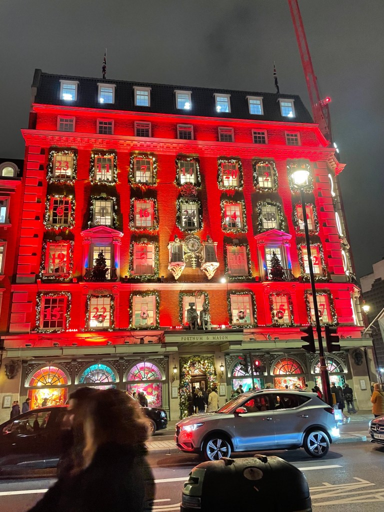 La facciata di Fortnum & Mason a Londra decorata per Natale, con luci rosse e ghirlande festive, illuminata da addobbi natalizi, mentre le persone si avviano verso l'ingresso.