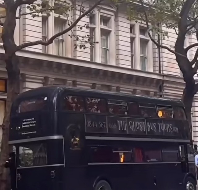 Un autobus nero per tour fantasma parcheggiato davanti a un edificio con finestre storiche e alberi visibili nei dintorni.
