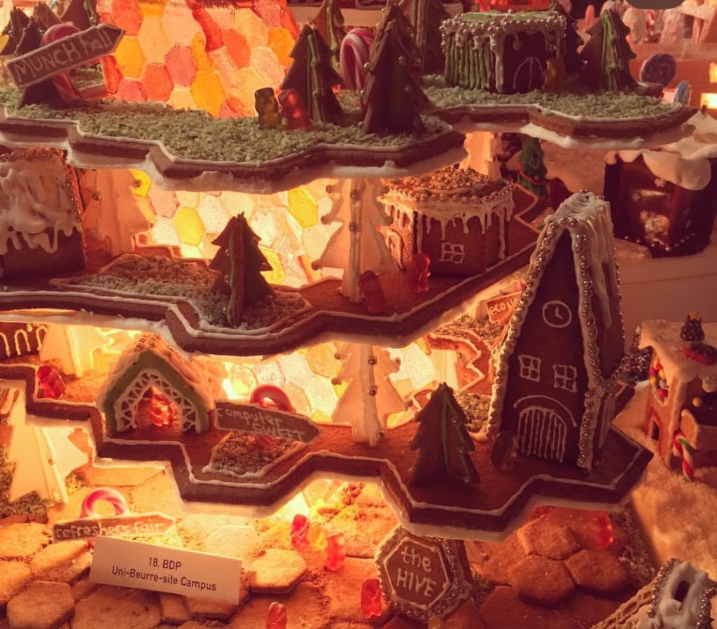 Struttura decorativa natalizia in biscotto di pan di zenzero, con alberi, case e decorazioni colorate, illuminata da luci calde.