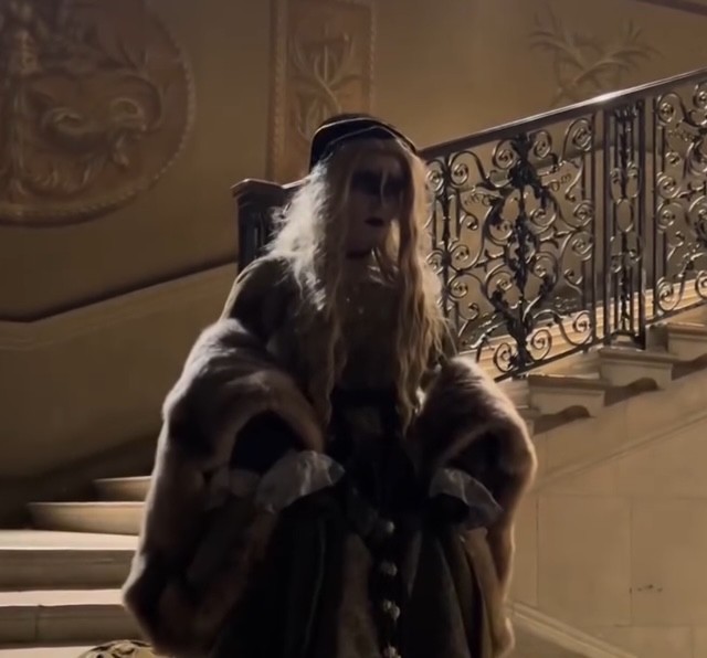 Un personaggio spettrale in costume di epoca, con capelli biondi e trucco inquietante, posizionato su una scala ornata di Hampton Court a Londra.