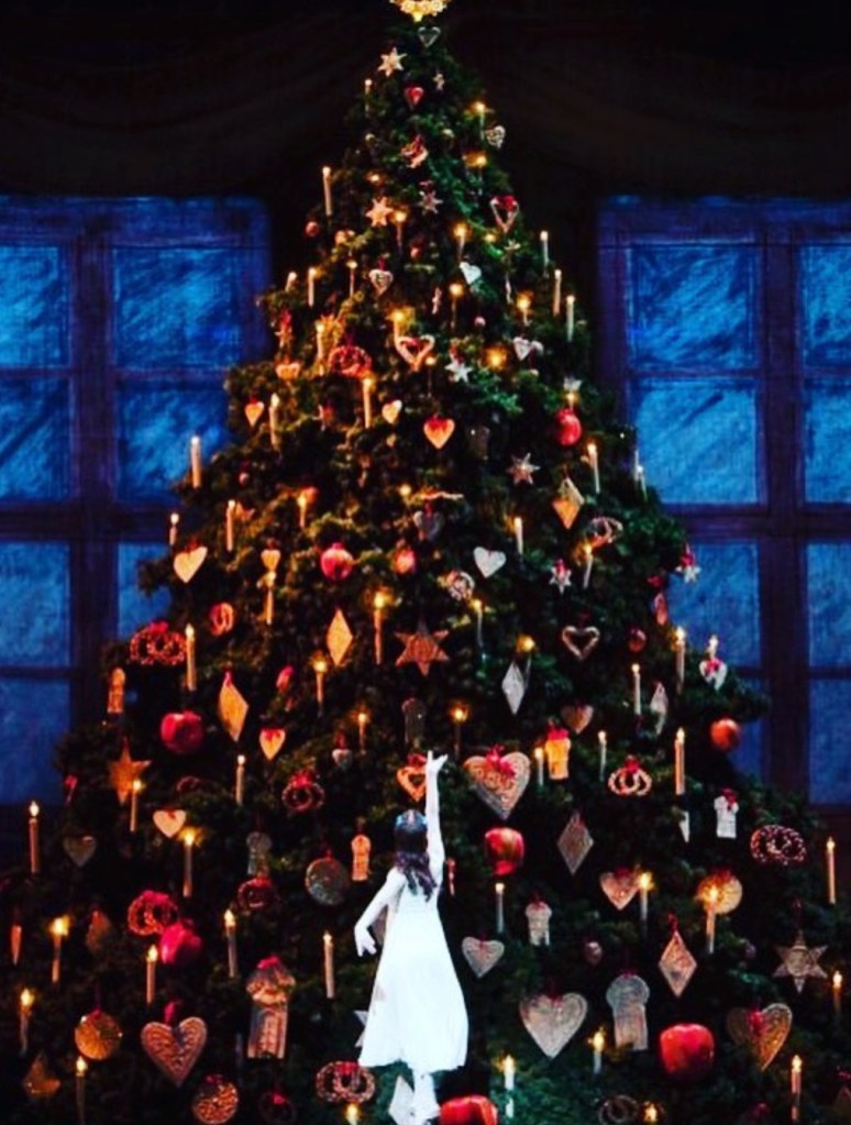 Un albero di Natale decorato con luci e ornamenti, accompagnato da una figura che cerca di toccarne la cima.