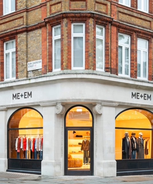 Esterno del negozio ME+EM situato su un angolo di un edificio a Londra, con finestre ampie e una selezione di abbigliamento esposto.