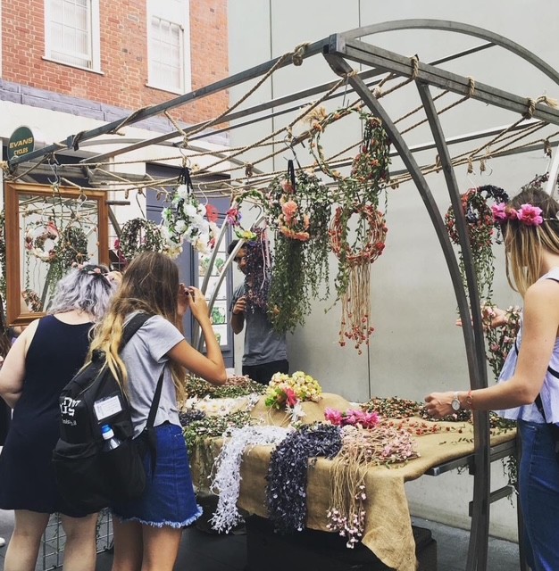 Due persone esplorano un banco di fiori in un mercato, circondate da ghirlande floreali e decorazioni naturali appese.