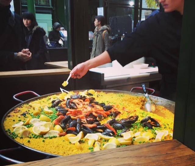 Uno chef che serve una grande paella con frutti di mare e limoni, mentre i visitatori del mercato osservano il piatto.