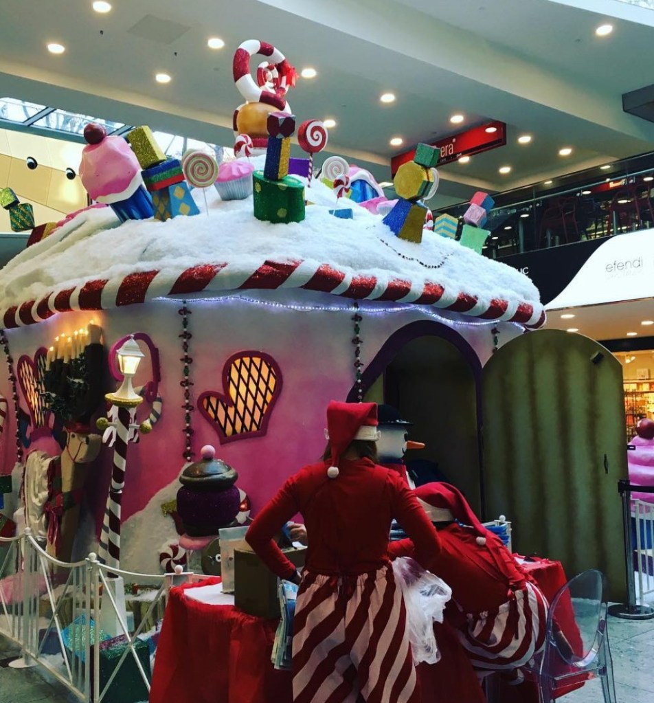 Un chiosco di dolci natalizi decorato con topping colorati, caramelle e regali, con due figure vestite di rosso che interagiscono davanti.