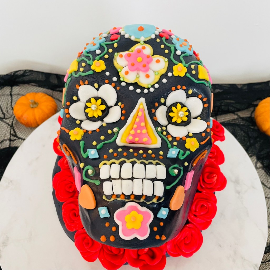 Torta decorata a forma di teschio messicano, colorata con fiori e dettagli vivaci, con zucche decorative sullo sfondo.