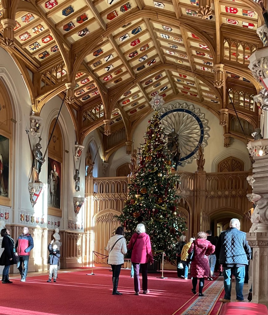 Interno di un castello decorato per Natale, con un grande albero addobbato e un soffitto in legno intagliato. Persone in visita osservano la bellezza dell'ambiente.