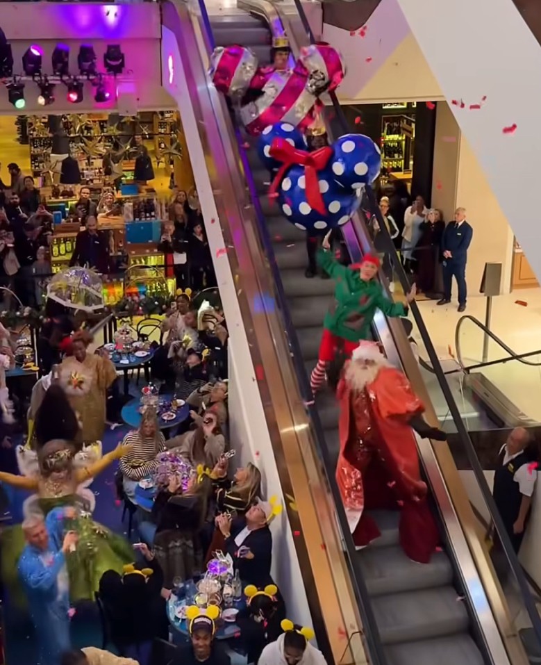 Un evento festivo da Selfridges a Londra con persone che si godono una performance su una scala mobile, caratterizzata da decorazioni natalizie e costumi colorati.