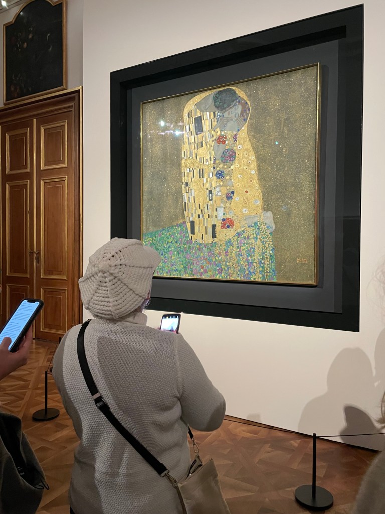 Una persona in maglione bianco e cappello di lana osserva un famoso dipinto incorniciato, il bacio di Klimt,  mentre utilizza un telefono per scattare una foto. Sullo sfondo, si intravedono pareti di legno e altre opere d'arte.
