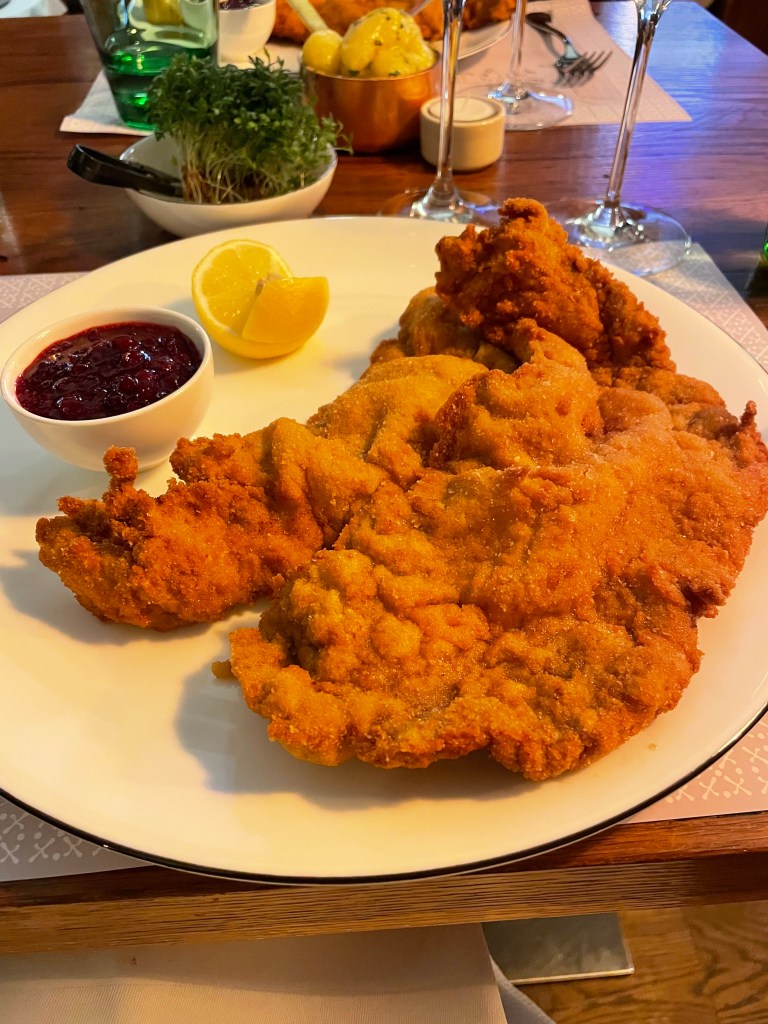 Una porzione di cotoletta viennese croccante servita con una fetta di limone e confettura di mirtilli rossi, accompagnata da contorni vari tra cui patate e insalata.