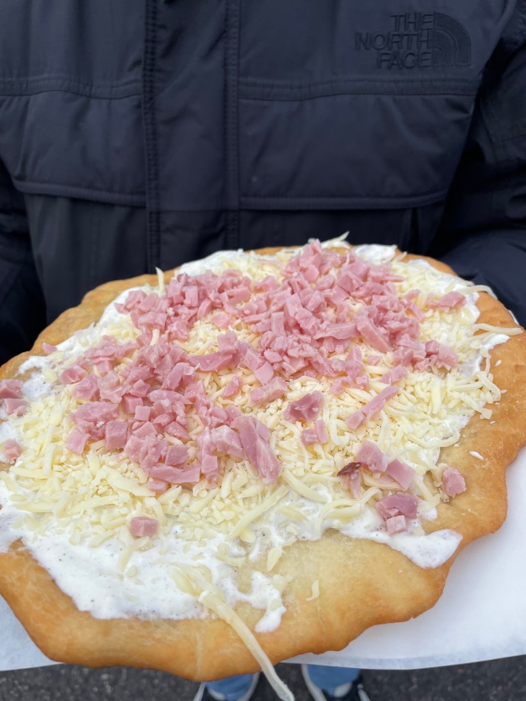 Frittella grande con panna acida, formaggio e prosciutto, tenuta in mano da una persona.