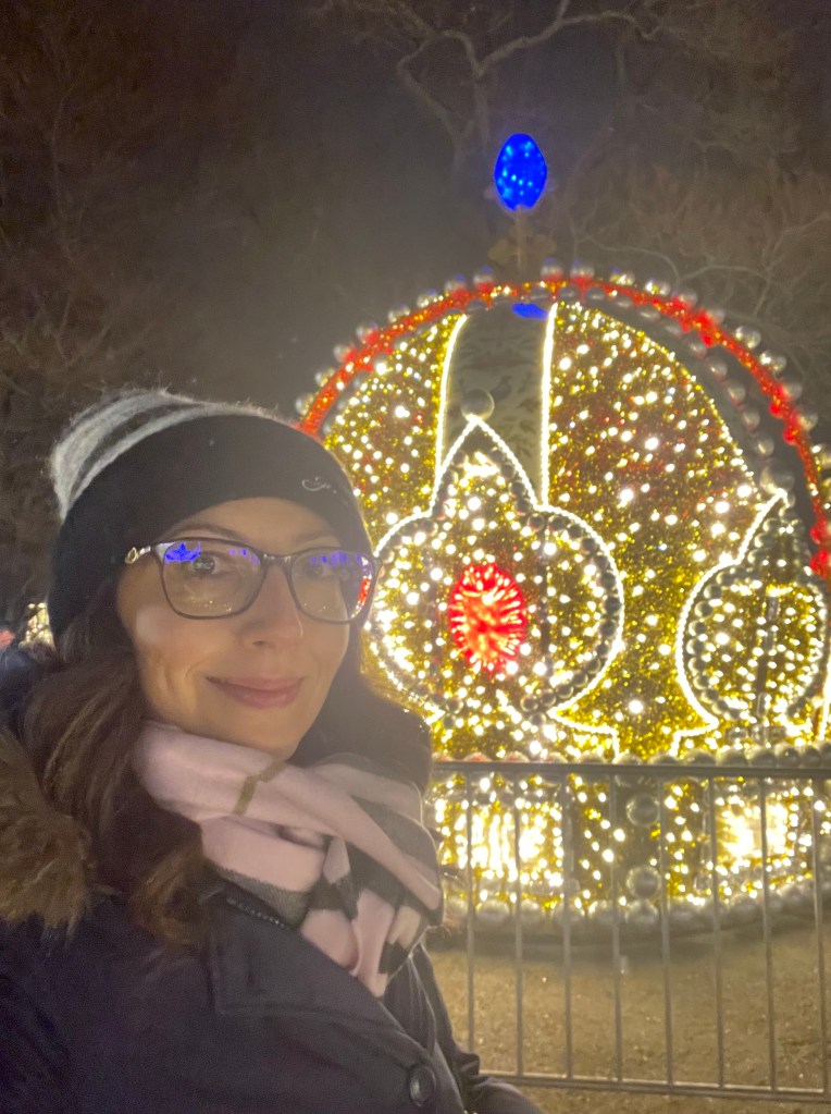 Una persona sorridente in primo piano, vestita con un cappello e una sciarpa, davanti a decorazioni natalizie luminose in un parco di Vienna di notte si tratta di Schloss Schonbrunn.