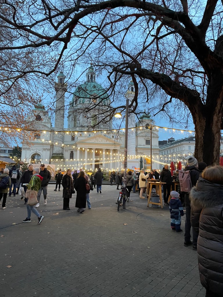 Mercatino di Natale a Vienna con illuminazioni festive e affollamento di visitatori, di fronte a un edificio storico con cupole verdi.