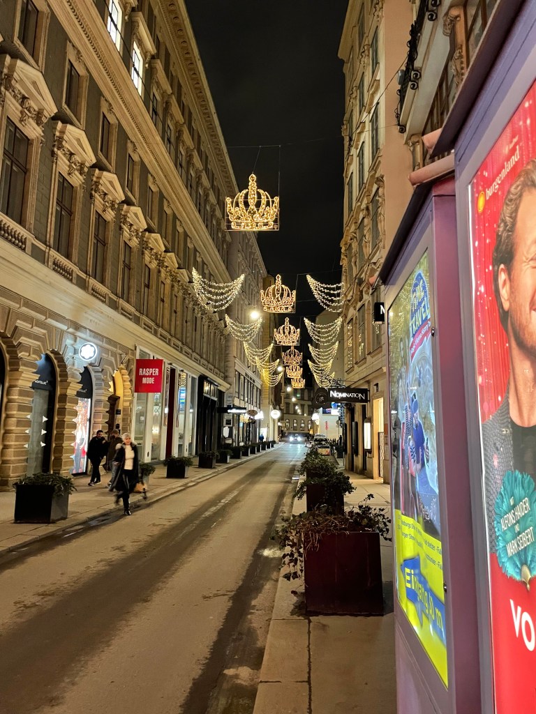 Una strada di Vienna illuminata da decorazioni natalizie a forma di corona, con negozi ai lati e pedoni che passeggiano, di sera.