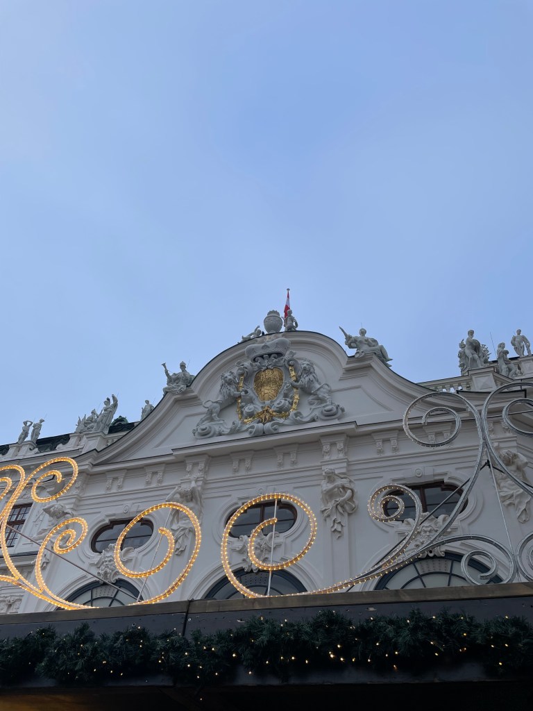 Dettaglio di un edificio storico di Vienna, ornato con decorazioni architettoniche e luci natalizie, contro un cielo azzurro.