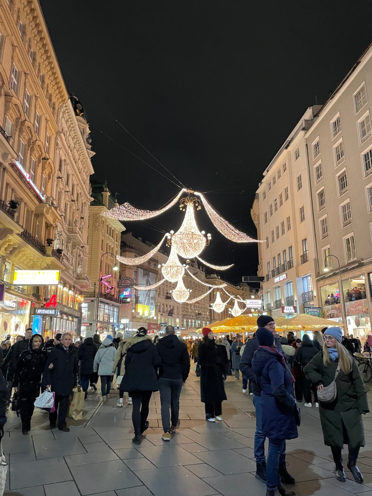 Una via di Vienna illuminata con decorazioni natalizie, lampadari di luci appesi e persone che passeggiano tra negozi e mercatini.