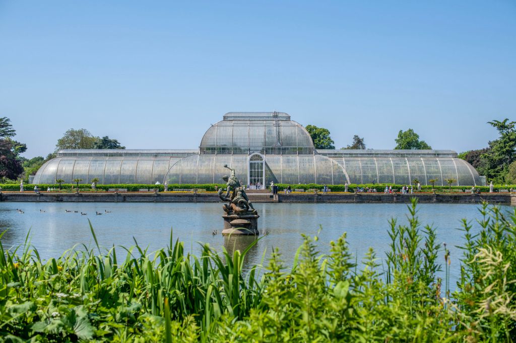 Una serra a Kew garden, la palm house, di vetro situata vicino a un lago, circondata da piante verdi e con visitatori in lontananza.