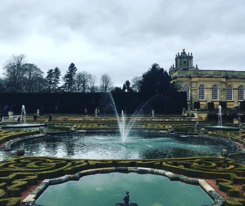 Vista di un giardino a Blenheim palace con una fontana centrale, circondato da aiuole decorative e alberi, in un giorno nuvoloso.