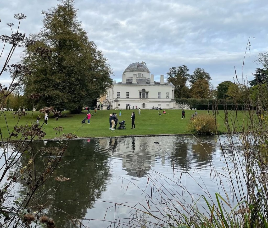Una vista della residenza storica di Chiswick a Londra, villa bianca con un giardino verde e persone che passeggiano, riflessa in un lago tranquillo.