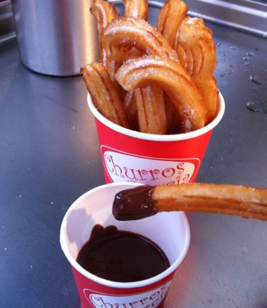 Due bicchieri rossi con churros, uno dei quali immerso in una ciotola di cioccolato fondente.