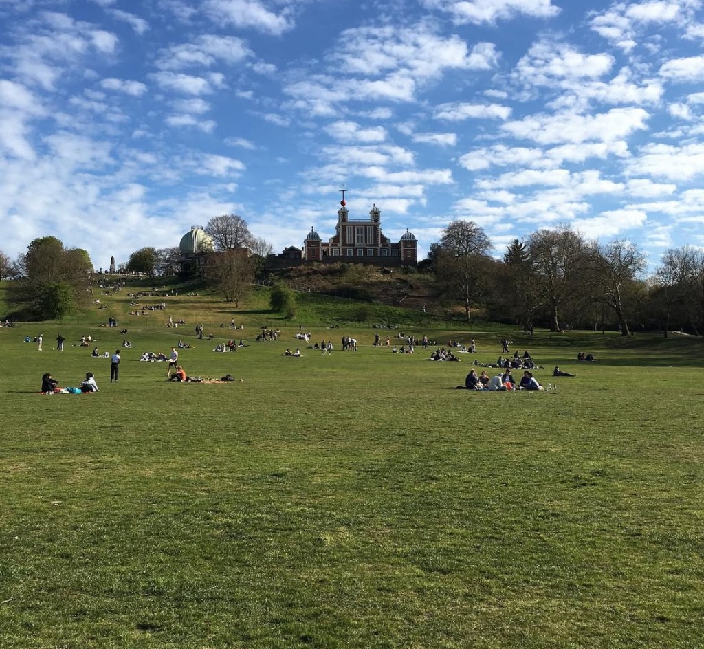 Vista de un grande parco di Greenwich a Londra con molte persone che si godono una giornata di sole, con l'Osservatorio sullo sfondo e un cielo parzialmente nuvoloso.
