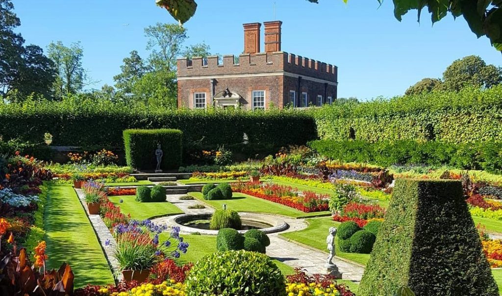 Giardino elegante di Hampton Court con aiuole fiorite e statue, sullo sfondo un edificio storico con torrette.