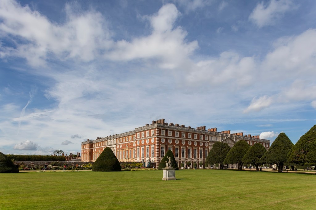 Vista del Palazzo di Hampton Court con giardini curati e alberi potati in un cielo sereno.