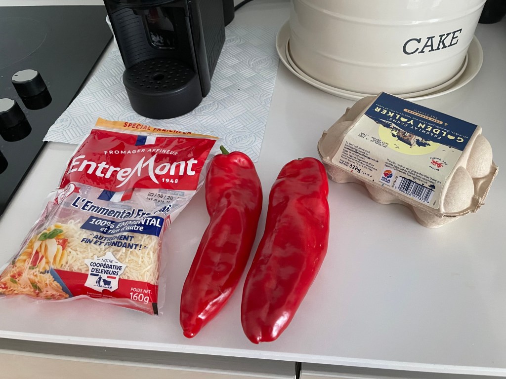 Una confezione di formaggio grattugiato, due peperoni rossi e un cartone di uova su un piano cucina con una caffettiera sullo sfondo. Pochi ingredienti per un piatto gustoso come una soffice frittata al forno.