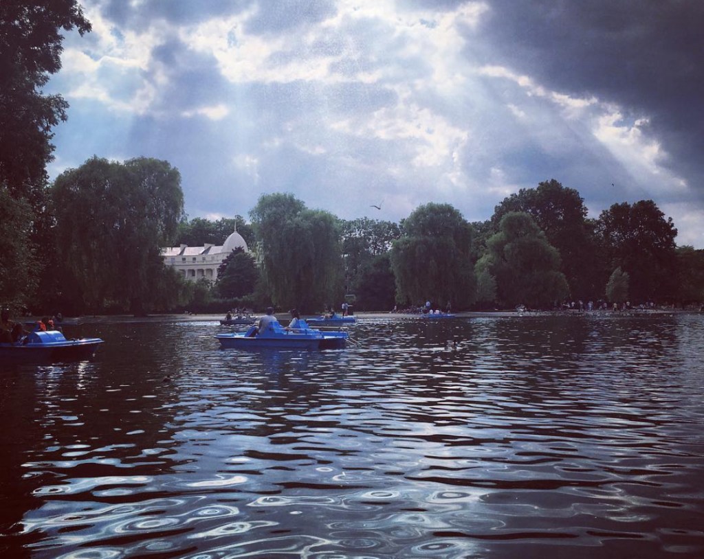 Un lago al Regent park di Londra  con persone su barche a remi blu, circondato da alberi verdi e un cielo nuvoloso.