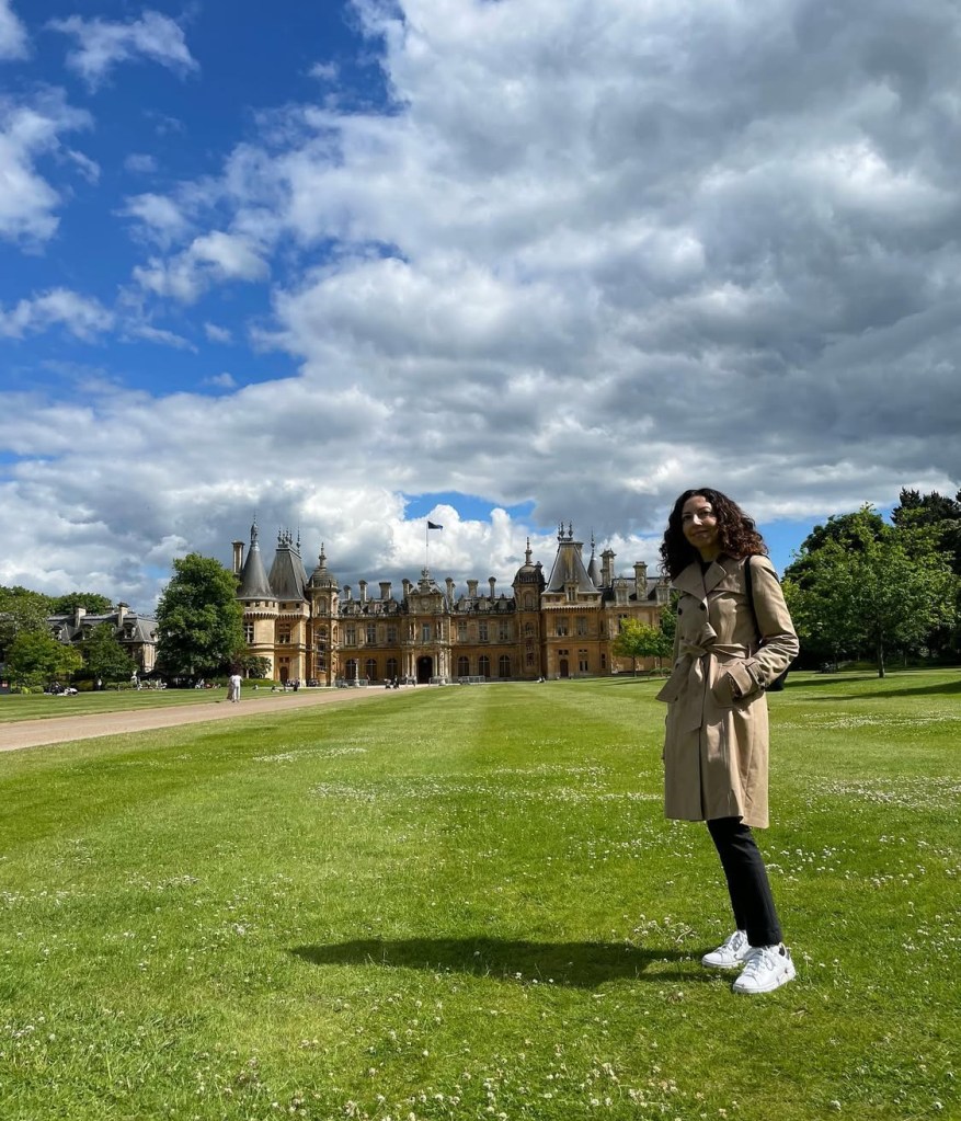 Io in un cappotto beige sorrido in un prato verde, con il grande palazzo storico di Waddeston manor sullo sfondo sotto un cielo nuvoloso.